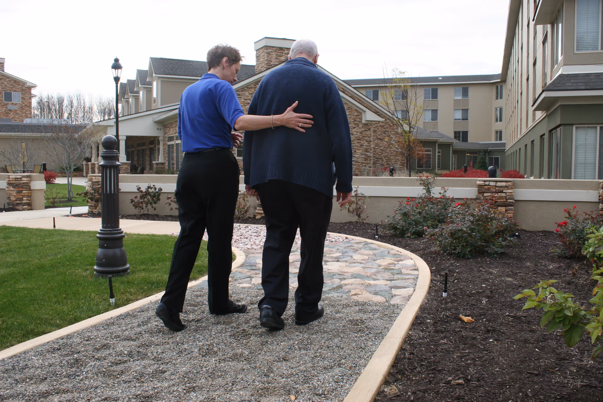 A caregiver in a blue shirt gently supporting an elderly man as they walk together on a stone and gravel pathway in the garden area of a senior living facility with multi-story buildings in the background.