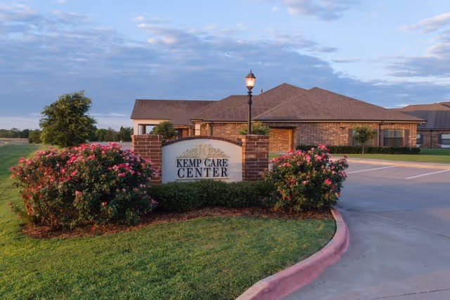 Exterior view of Kemp Care Center building with a brick facade, a sign displaying the facility name surrounded by bushes with pink flowers, a lamp post, and a paved driveway under a partly cloudy sky.