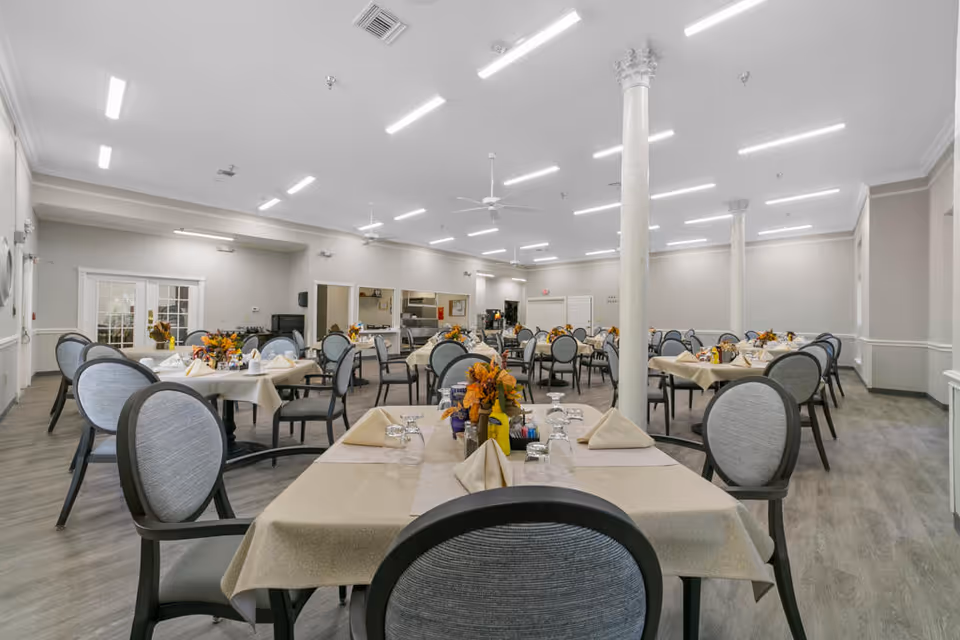 A spacious dining room with multiple round tables covered with beige tablecloths, each set with folded napkins, glasses, and floral centerpieces. The room has light gray walls, white ceiling with bright linear lights, ceiling fans, and several white columns. Chairs with gray cushioned backs and seats surround the tables. The far end of the room has a serving area with kitchen equipment visible.