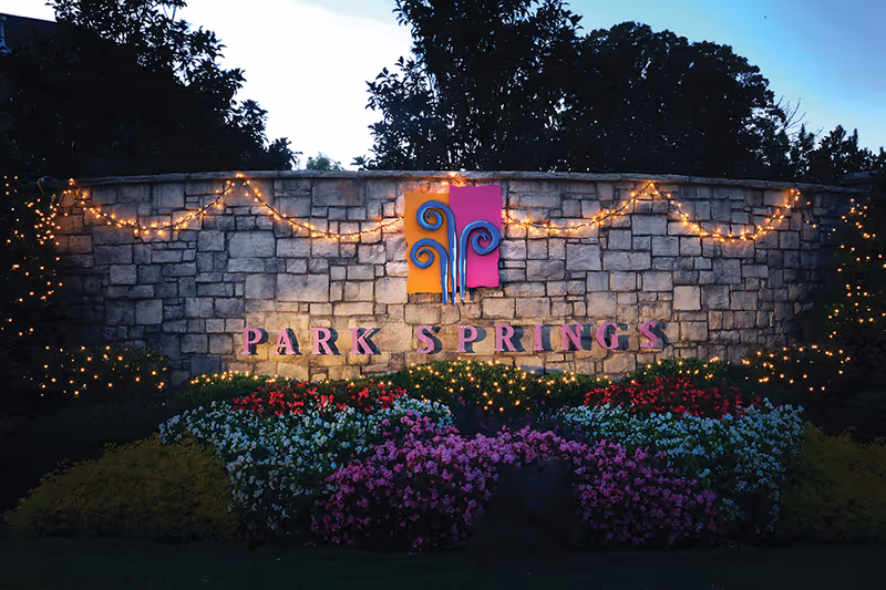 Stone wall with the sign 'PARK SPRINGS' illuminated by string lights, surrounded by colorful flowers and greenery at dusk.