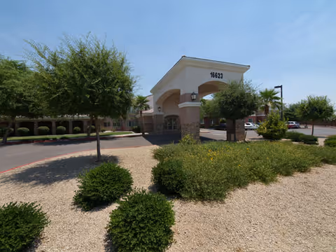 Exterior view of Chaparral Winds Assisted Living facility entrance with a covered drop-off area, surrounded by desert landscaping including bushes, small trees, and gravel. The building number 16623 is visible above the entrance.