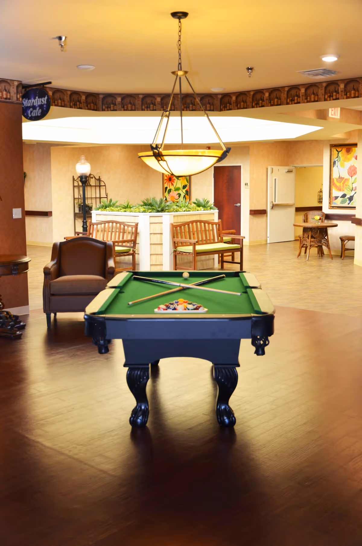 Interior of a senior living facility common area featuring a pool table with cues and balls set up, surrounded by wooden benches with green cushions, a brown armchair, and a hanging light fixture above the pool table. The room has warm lighting, wooden flooring, and decorative plants in the background. There is a sign that reads 'Stardust Cafe' hanging from the ceiling and colorful floral artwork on the walls.