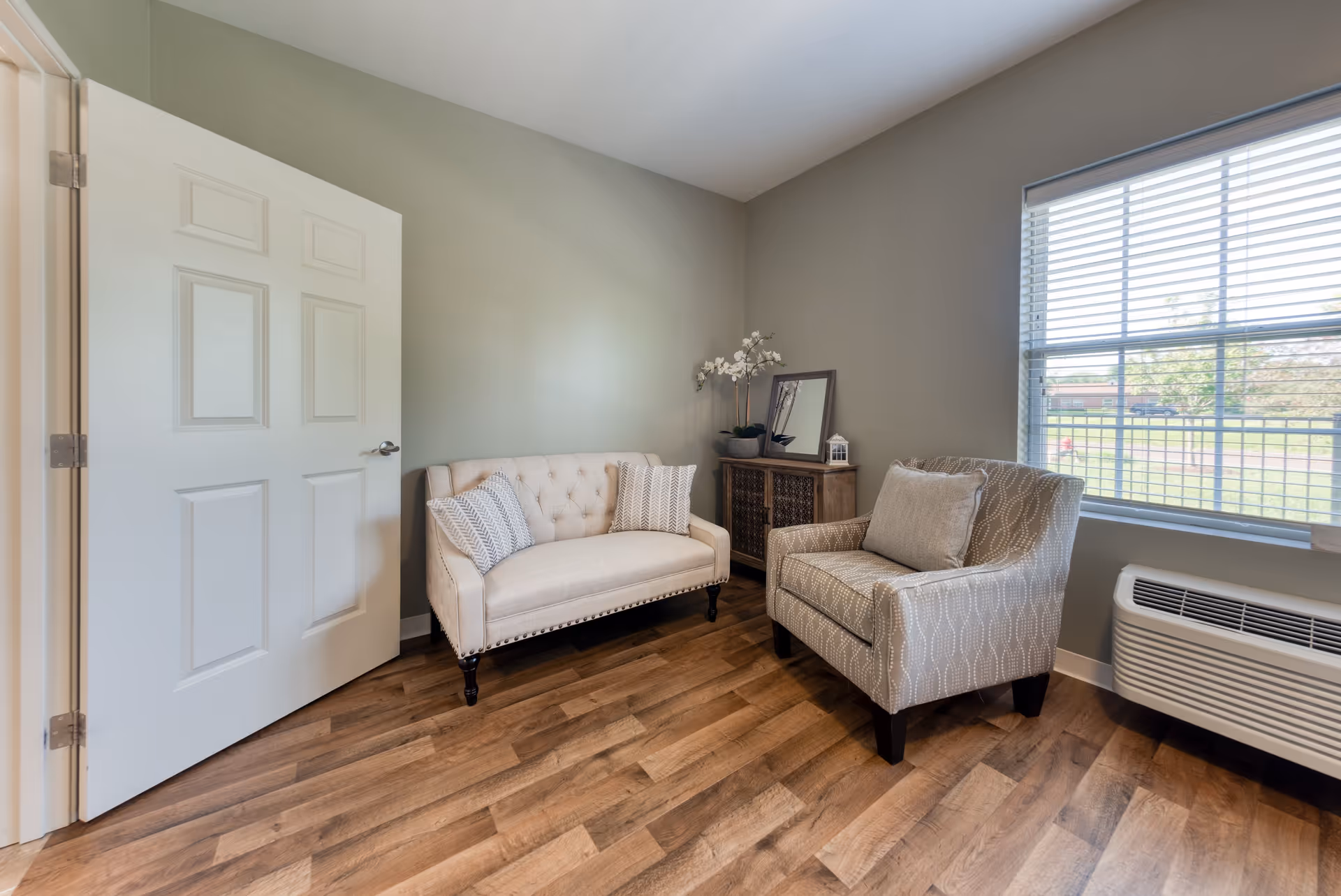 A small sitting area with a beige tufted loveseat and a patterned armchair with a cushion. There is a wooden cabinet with decorative items including a potted orchid, a mirror, and a small lantern. The room has light gray walls, wood flooring, a white door, and a window with blinds showing an outdoor view.