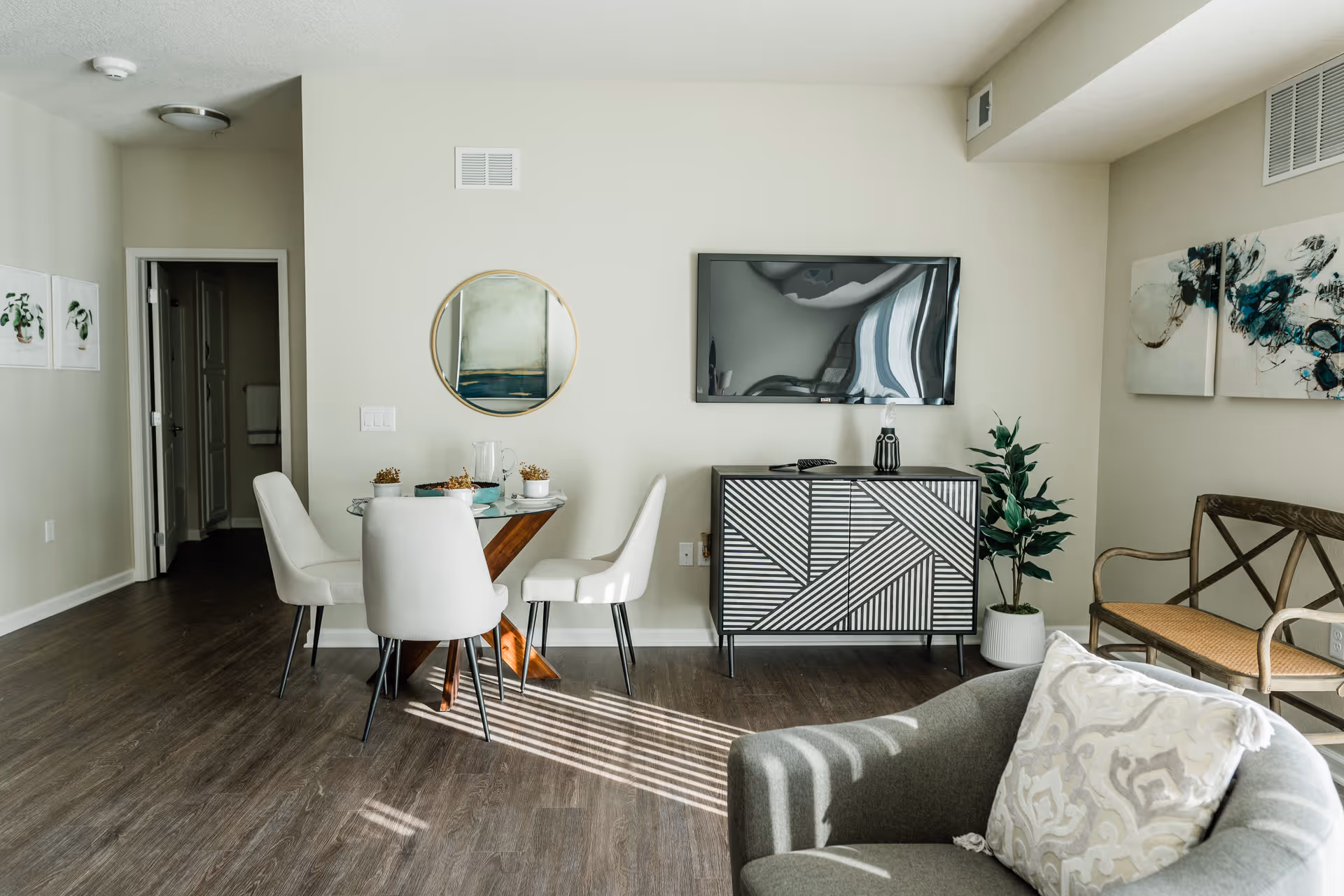 A modern living and dining area with a round glass dining table surrounded by four white chairs. A flat-screen TV is mounted on the wall above a black and white patterned cabinet. There is a potted plant next to the cabinet and a wooden bench with a woven seat against the wall. A gray armchair with a patterned cushion is in the foreground. The room has light-colored walls, wood flooring, and decorative artwork on the walls.