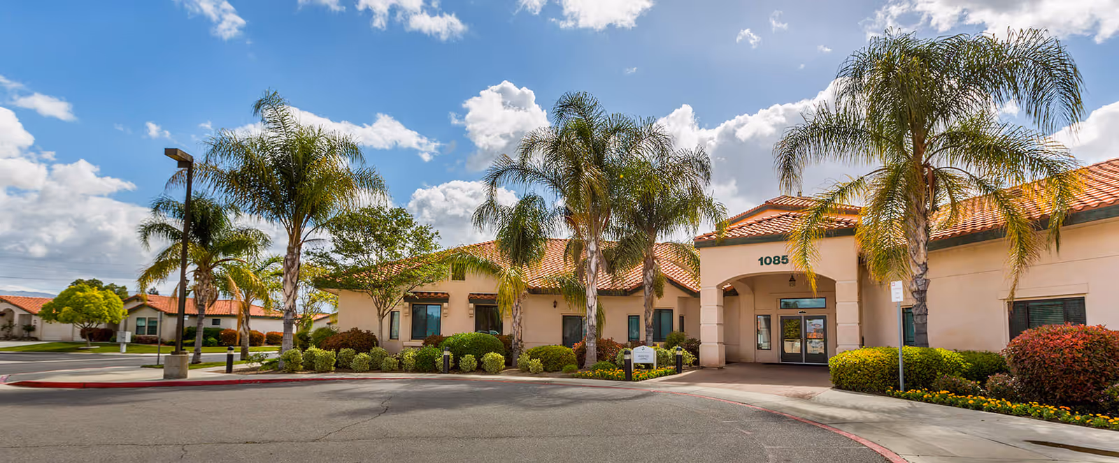 Exterior view of a senior living facility building with a beige facade and red tile roof, surrounded by palm trees and landscaped bushes under a partly cloudy blue sky. The building entrance is visible with the number 1085 above it.
