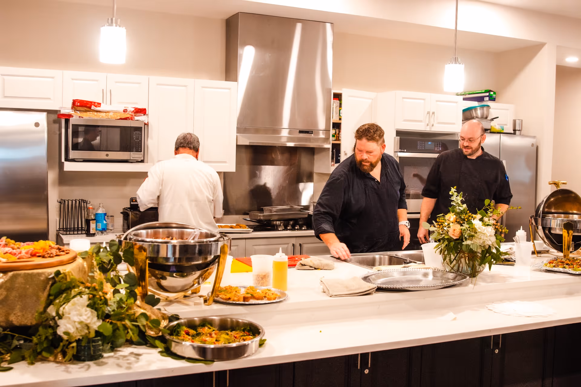 Three men working in a modern kitchen with white cabinets and stainless steel appliances. The kitchen island is decorated with flowers and various dishes of food. Two men are facing the counter, one is facing away, and the atmosphere appears busy and professional.