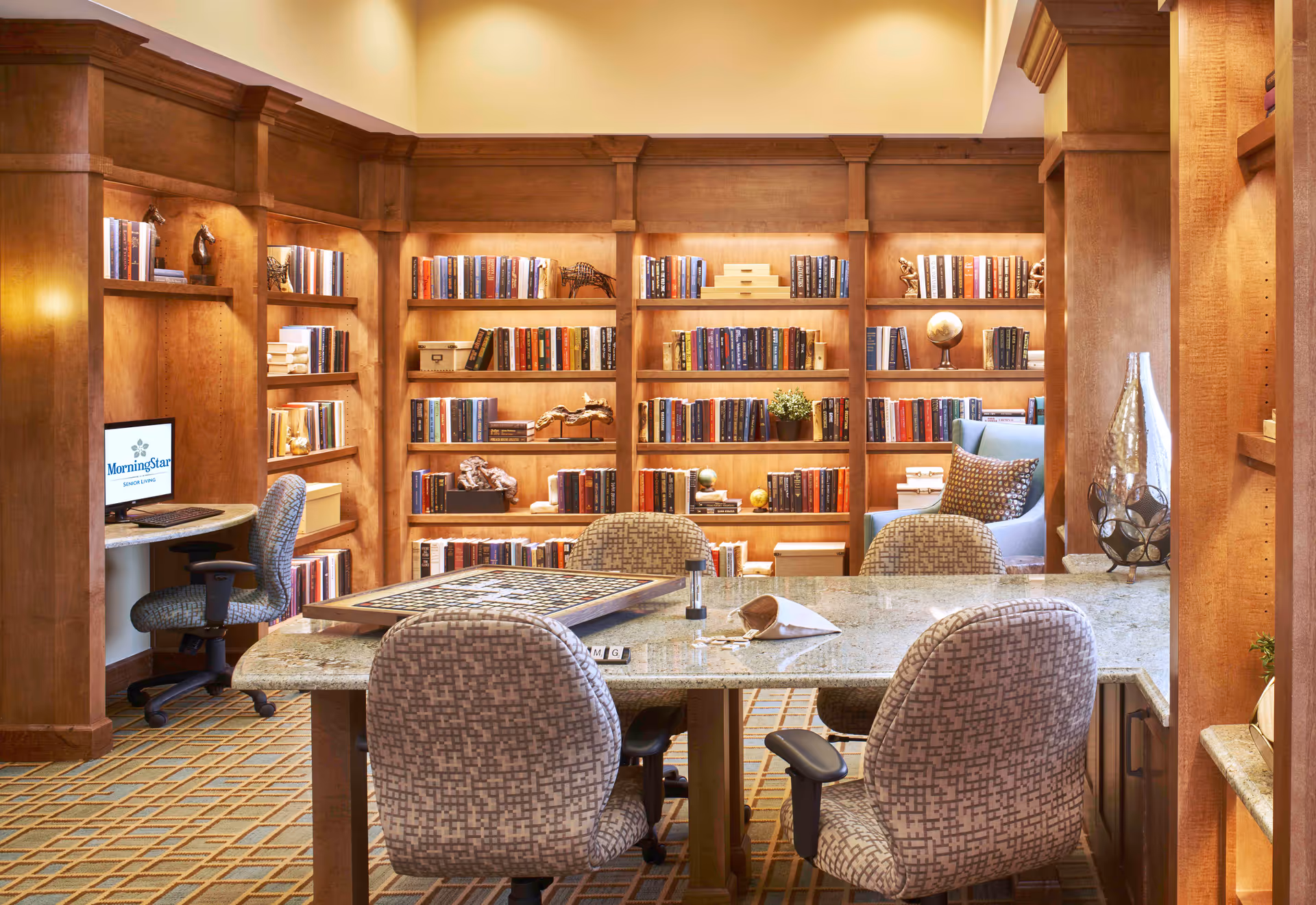 A cozy library or reading room with wooden bookshelves filled with books and decorative items. There is a large table with four patterned chairs around it, and a computer workstation with a chair on the left side. The room has warm lighting and a comfortable armchair with a pillow near the bookshelves.