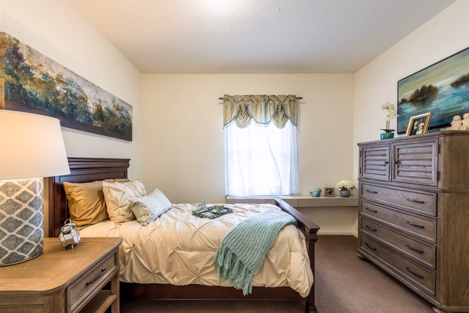 A cozy bedroom in Fairborn Assisted Living featuring a wooden bed with cream-colored bedding and decorative pillows. A light blue throw blanket is draped over the bed. Next to the bed is a wooden nightstand with a patterned lamp and a small clock. Across from the bed is a wooden dresser with framed pictures, a small plant, and decorative items on top. A window with sheer white curtains and a valance lets in natural light. A large landscape painting hangs above the bed, and another painting is above the dresser.