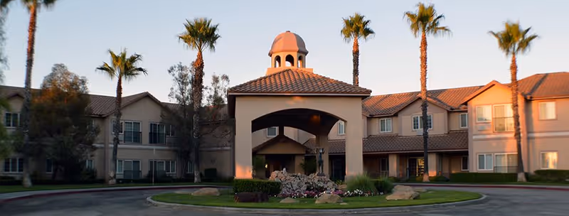 Exterior view of The Lakes Senior Living facility featuring a central covered entrance with a tiled roof and a small dome on top, surrounded by palm trees and a landscaped circular driveway.