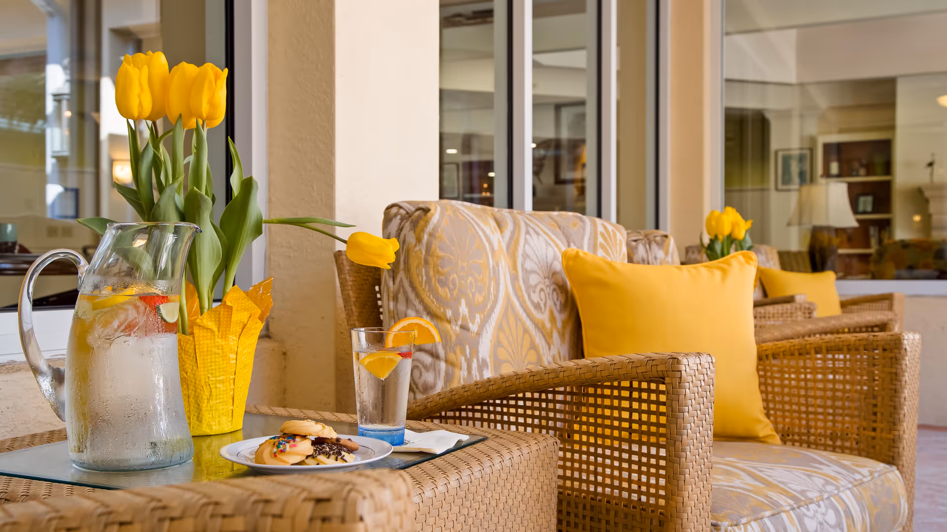 A cozy seating area with wicker chairs featuring patterned cushions and yellow pillows. On the glass-top table in front, there is a pitcher of water with lemon and cucumber slices, a glass of water with a lemon wedge and cherry, a plate of cookies, and a yellow potted plant with tulips. The setting appears to be a covered patio or sunroom with large windows showing an interior space in the background.