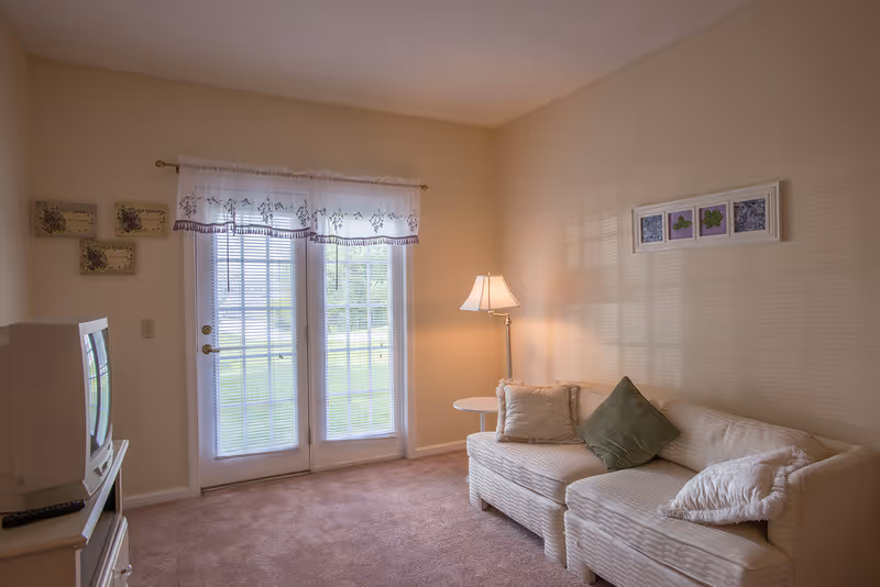A cozy living room with a white couch adorned with three pillows, a floor lamp beside it, and a small round side table. There are French doors with sheer white curtains letting in natural light. A TV sits on a white stand to the left, and framed artwork is hung on the walls.