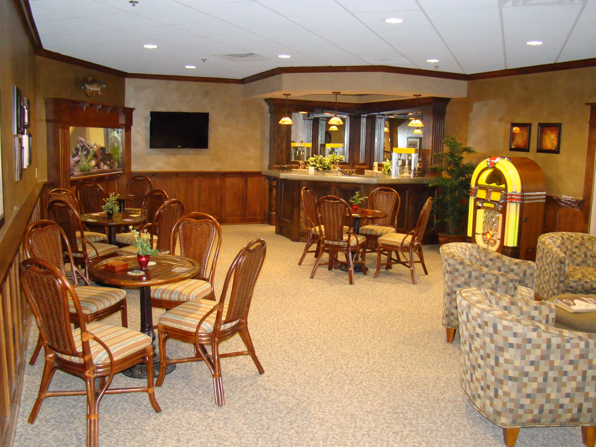 A cozy common area with multiple round tables and wooden chairs with striped cushions, a bar counter with hanging pendant lights, a wall-mounted flat screen TV, an aquarium built into the wall, a jukebox, and several patterned armchairs arranged around a small table.
