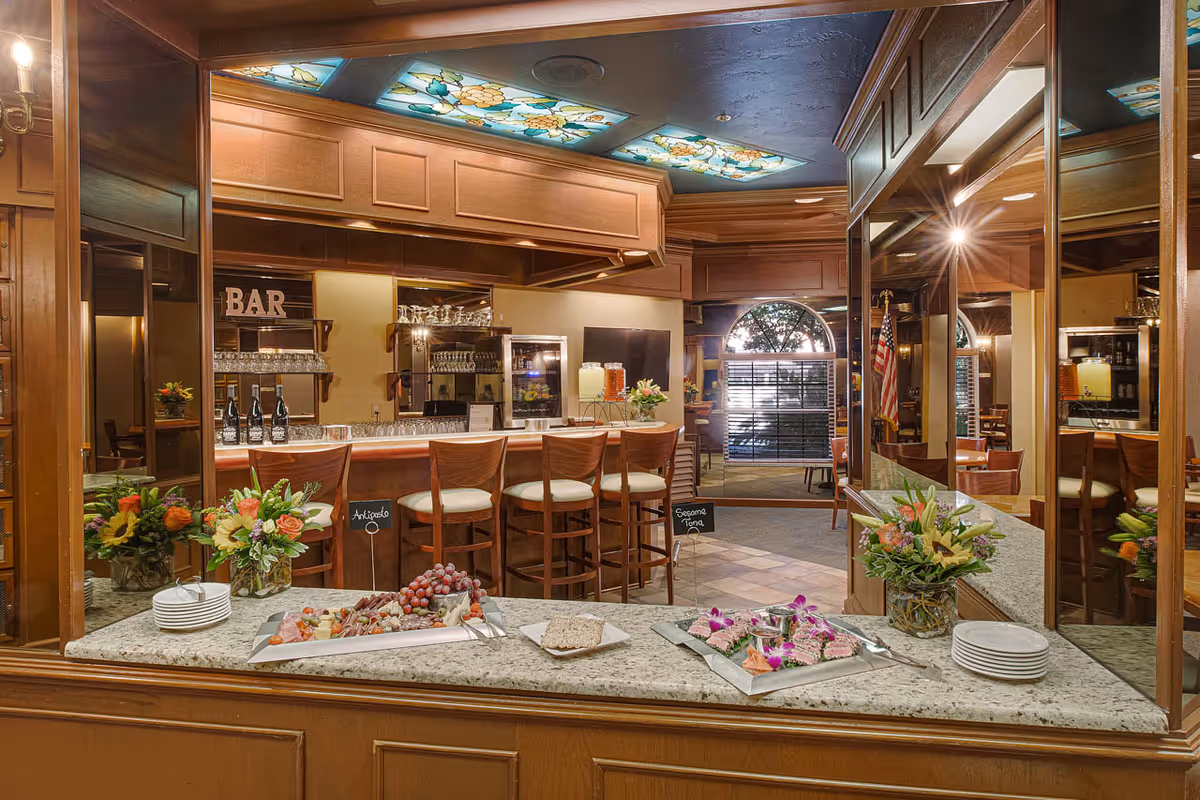 Interior view of a bar area in a senior living facility featuring a granite countertop with platters of appetizers and flowers. Behind the counter are wooden bar stools, glassware, and beverage dispensers. The ceiling has decorative stained glass panels, and there is an American flag in the background near an arched window with blinds.