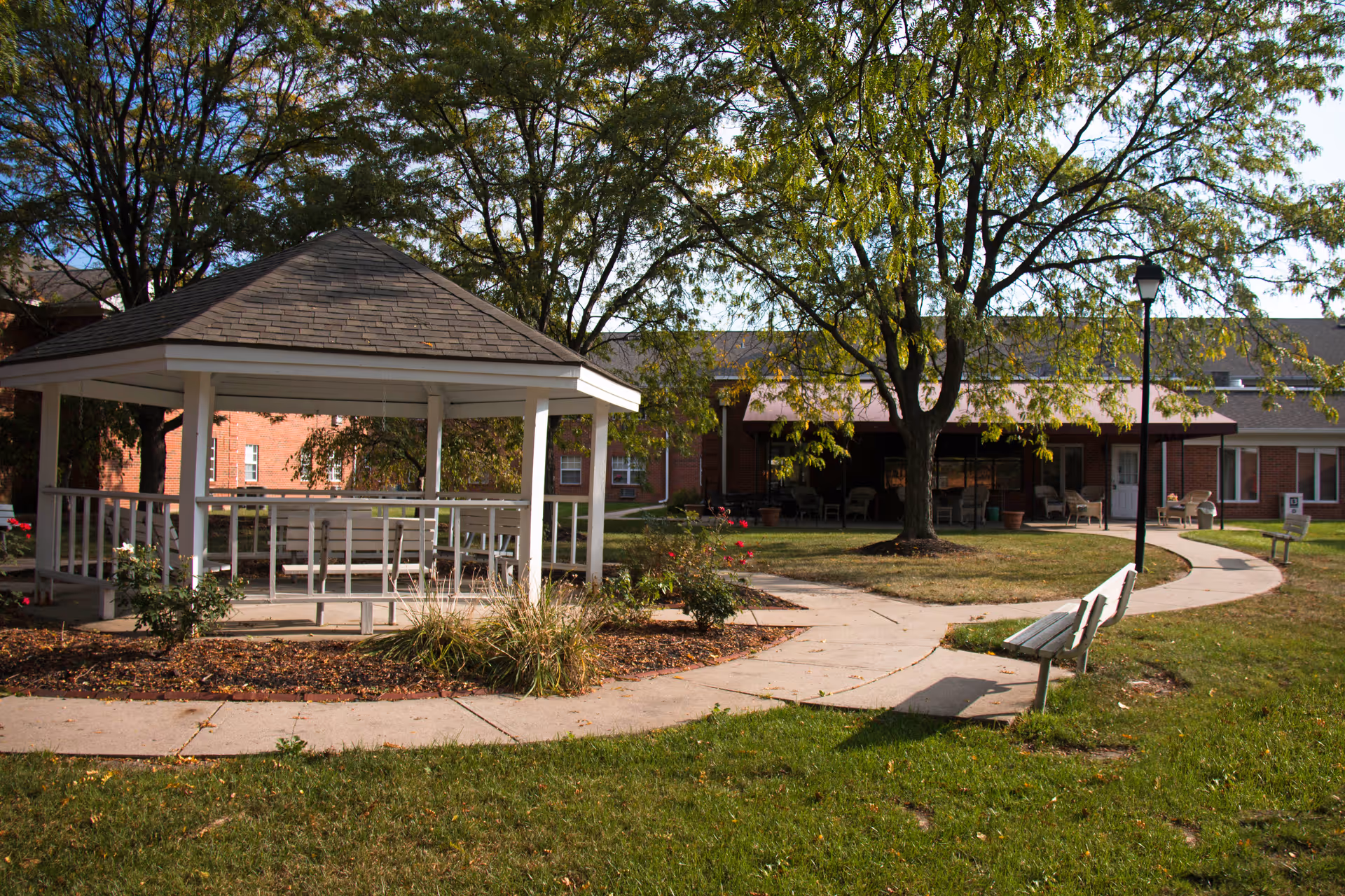 Outdoor area of Wyndmoor of Castleton featuring a white gazebo with benches inside, surrounded by trees, flower beds, and a curved concrete walkway. There are benches along the walkway and a brick building with a covered patio in the background.