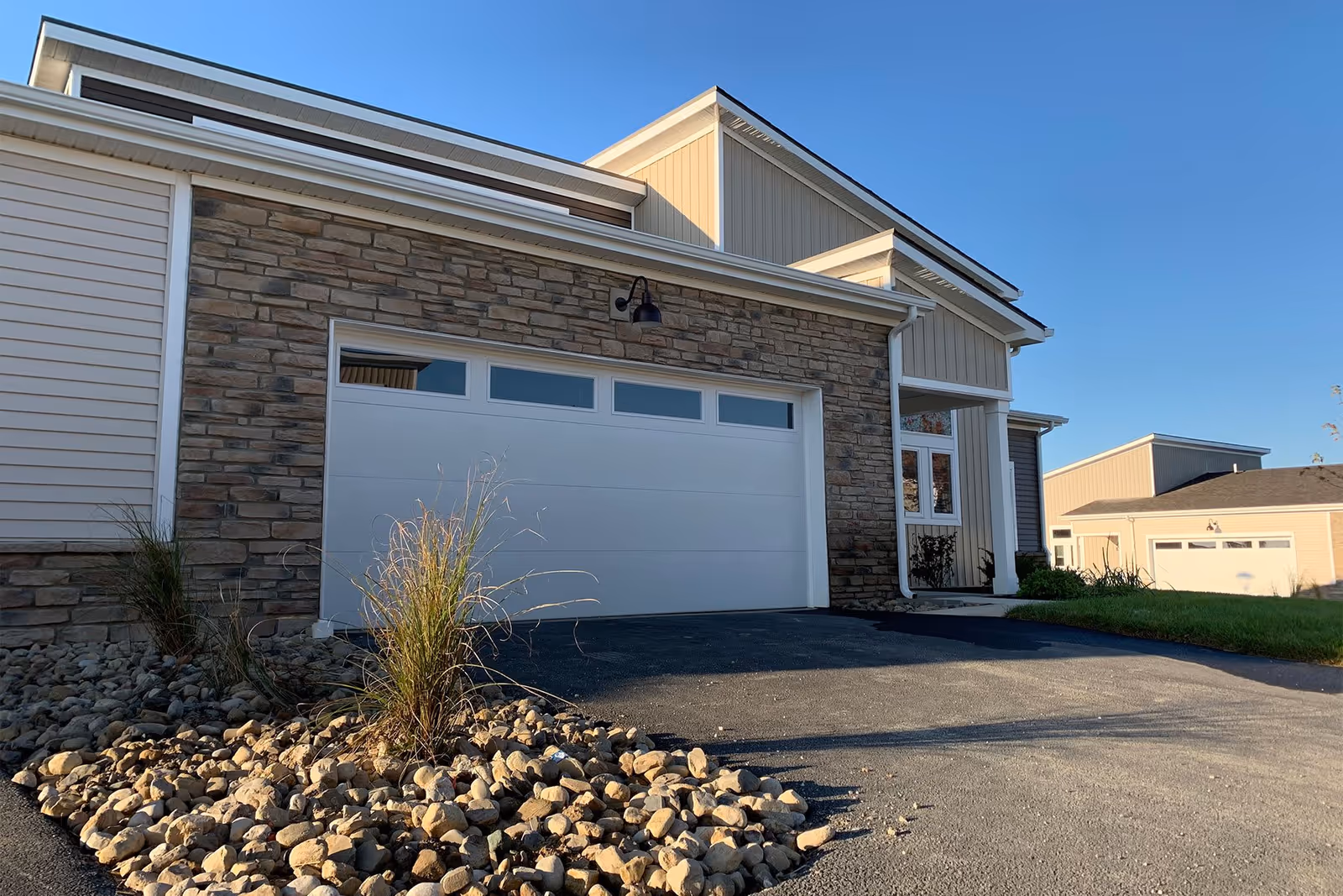 Exterior view of a modern residential building with a stone facade and a white garage door. There is a small landscaped area with rocks and ornamental grass in front of the garage. Another similar building is visible in the background under a clear blue sky.
