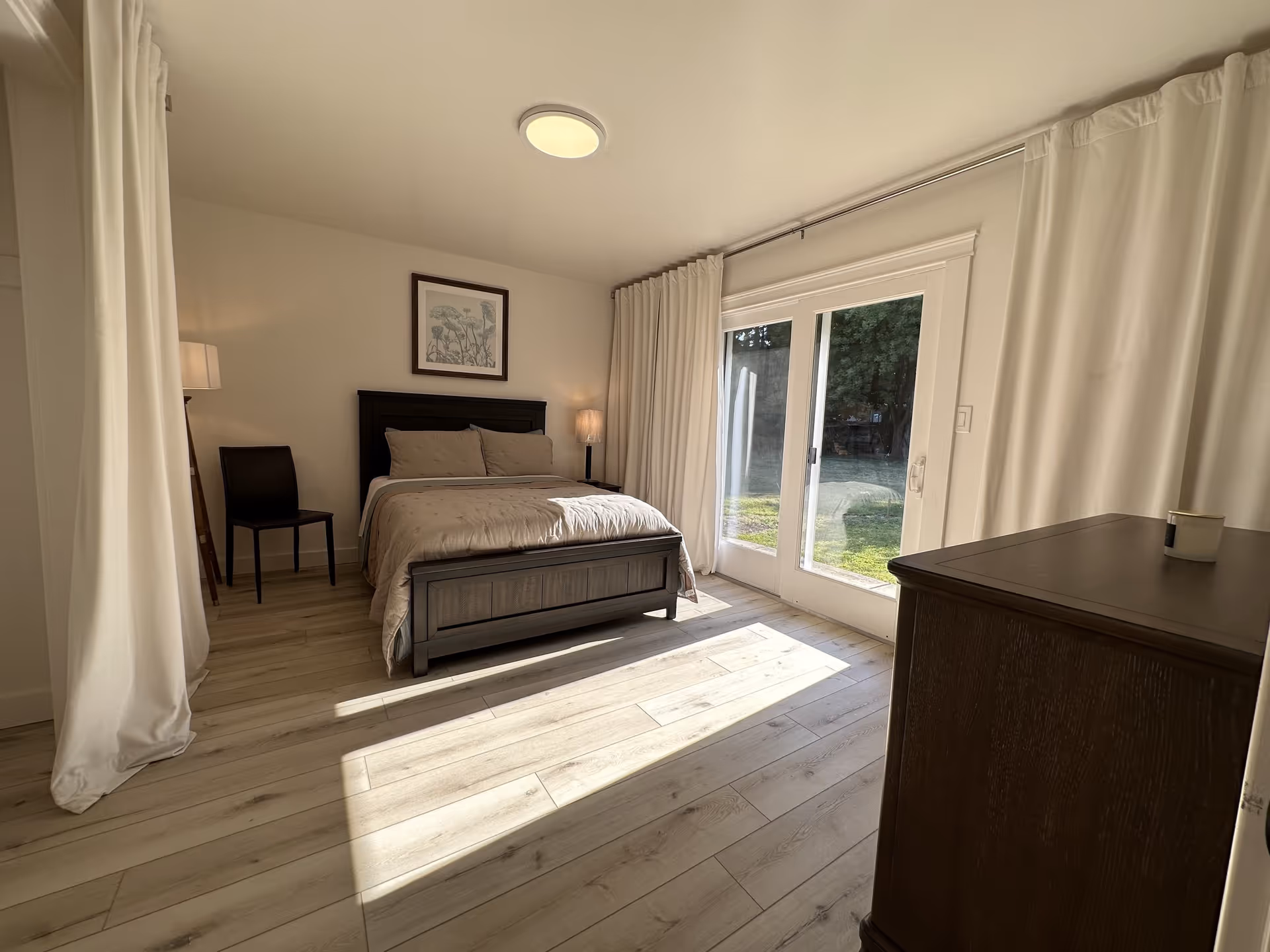 A bright bedroom with light wood flooring, a dark wooden bed with beige bedding, a black chair, a floor lamp, a framed picture above the bed, and large glass sliding doors with white curtains letting in natural light and showing a green outdoor area.