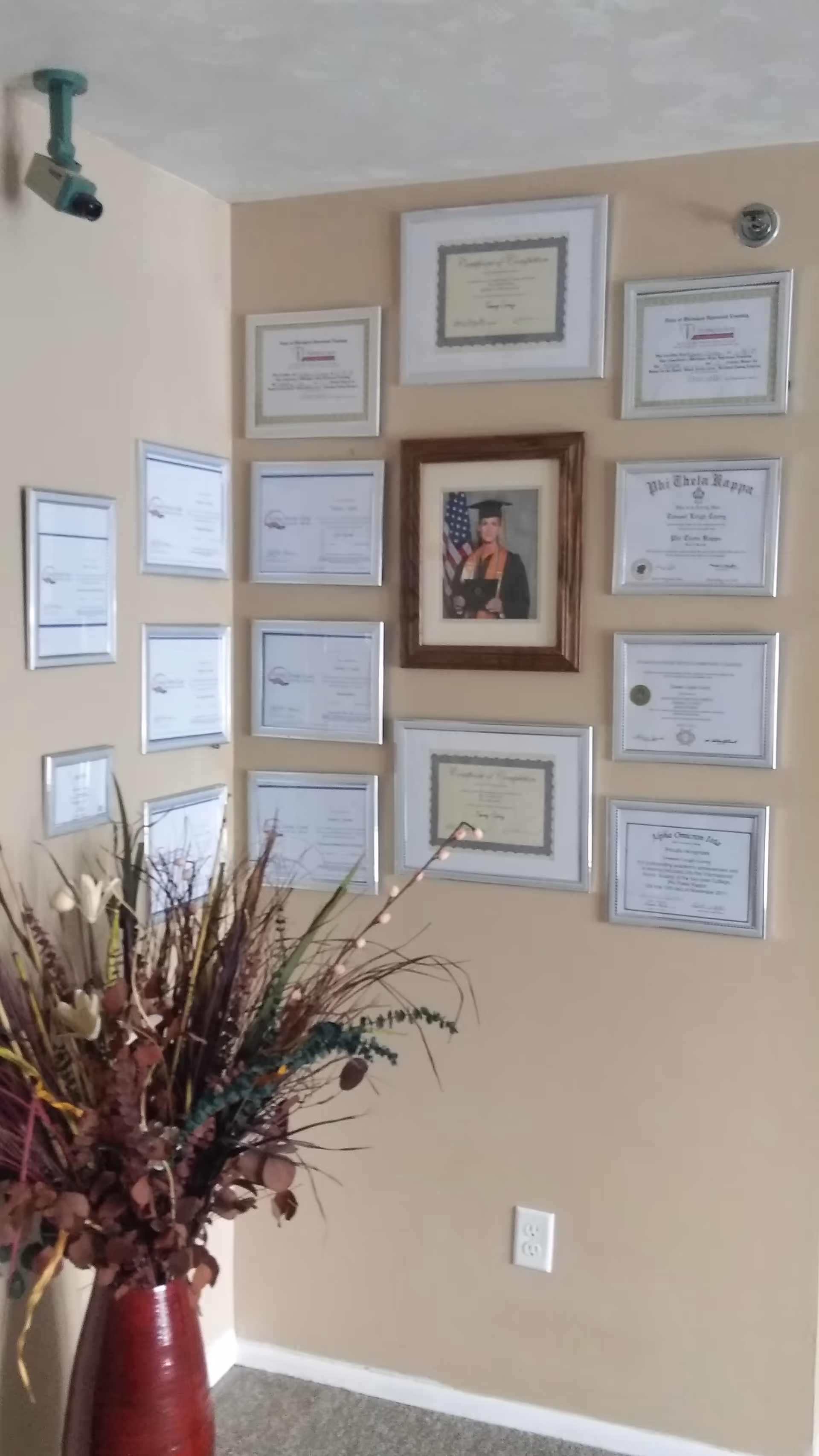 A beige wall decorated with multiple framed certificates and a central framed graduation photo. Below the frames is a red vase filled with dried flowers and branches. A security camera is mounted on the ceiling in the upper left corner.