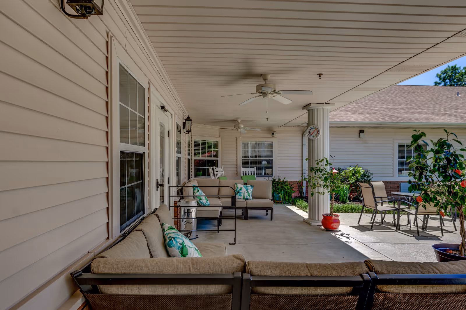Covered outdoor patio area at Garrett Woods Senior Living Community with cushioned seating, decorative pillows, ceiling fans, potted plants, and a small table with chairs. The patio is adjacent to a building with beige siding and multiple windows.