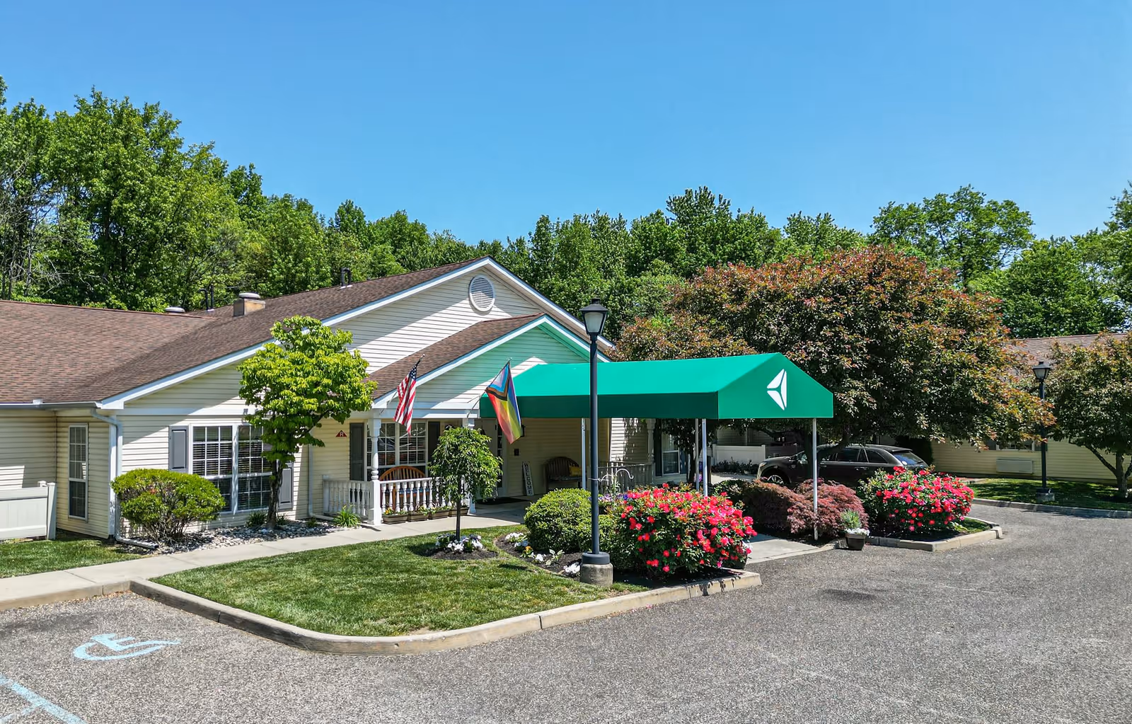 Exterior view of Arden Courts - ProMedica Memory Care Community in Cherry Hill, showing a single-story building with beige siding and a brown roof. There is a green canopy over the entrance with a white logo, two flags displayed near the entrance, well-maintained landscaping with bushes and flowers, a parking area with a handicapped parking space, and trees in the background under a clear blue sky.