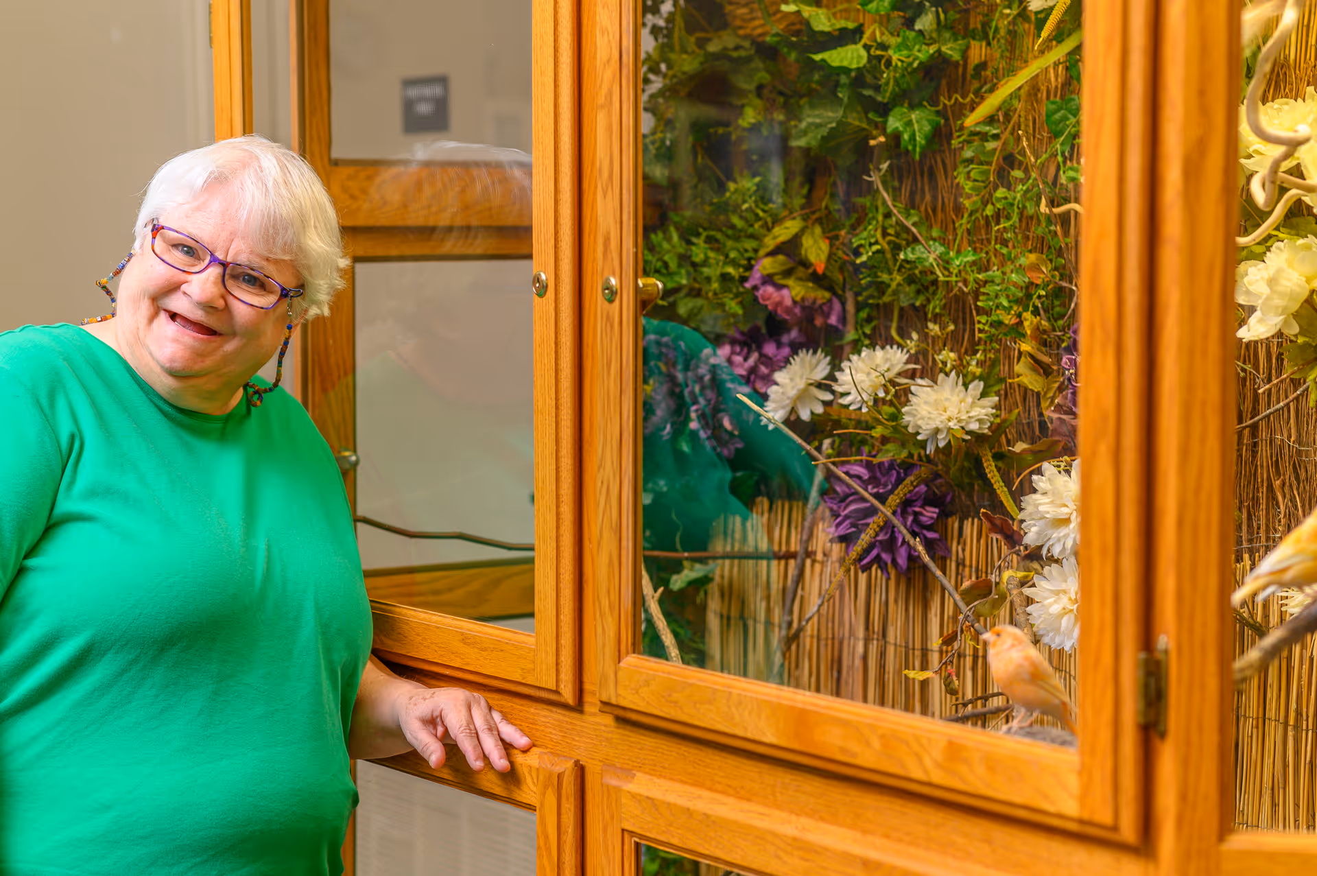An elderly woman wearing a green shirt and glasses stands smiling next to a wooden display cabinet filled with artificial flowers and decorative birds.