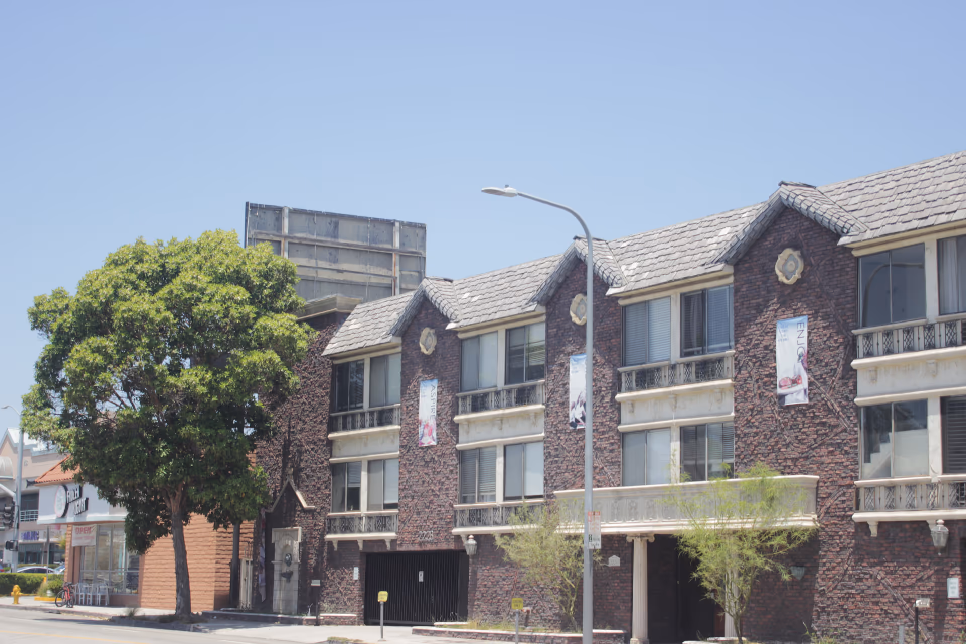 Exterior view of a three-story brick building with multiple windows and decorative elements, a large tree on the left side, and a streetlamp in front under a clear blue sky.