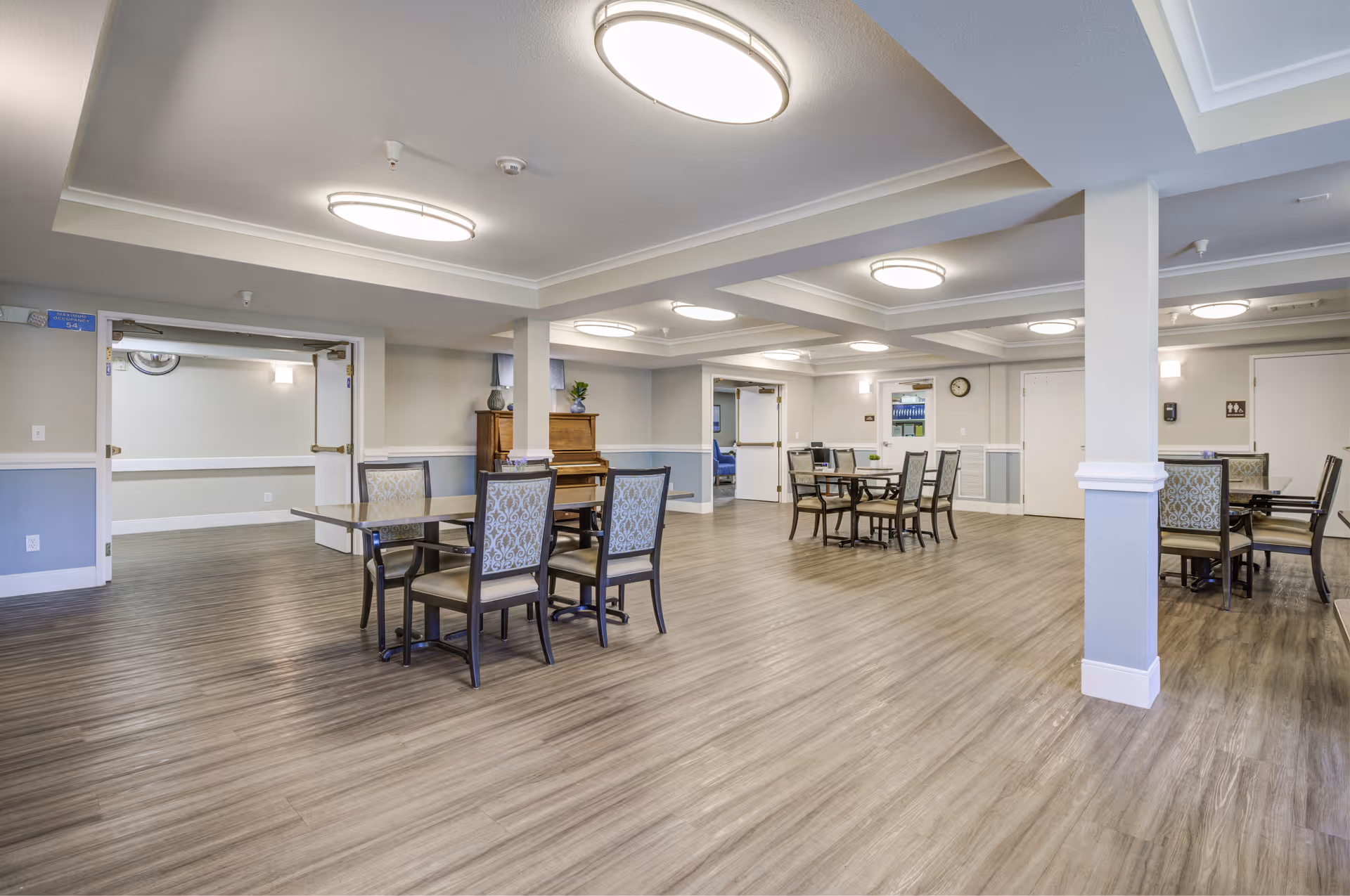 A spacious, well-lit common area with multiple tables and chairs arranged for seating. The room features wood-style flooring, light-colored walls with a blue accent, and ceiling lights. A piano is visible against one wall, and there are double doors leading to adjacent rooms.