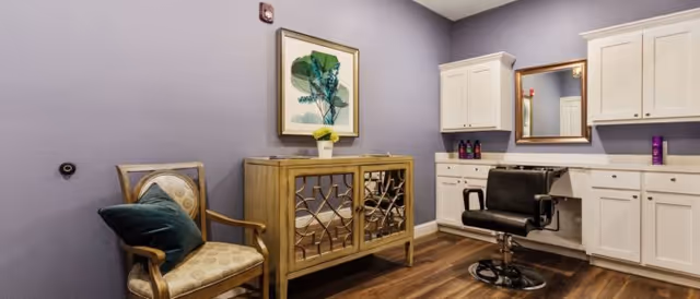 A small room with lavender walls featuring a wooden chair with a green cushion, a decorative wooden cabinet with a framed botanical print above it, and a black salon-style chair in front of a mirror mounted on white cabinetry with countertop and storage.