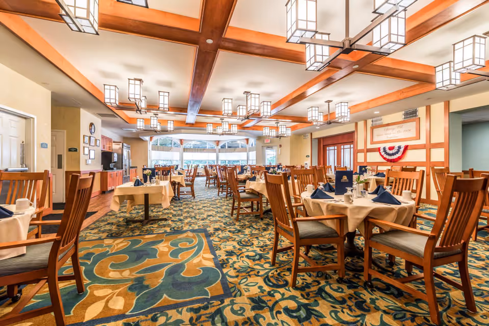 Spacious dining room with multiple set tables and wooden chairs on a patterned carpet under decorative ceiling lights.