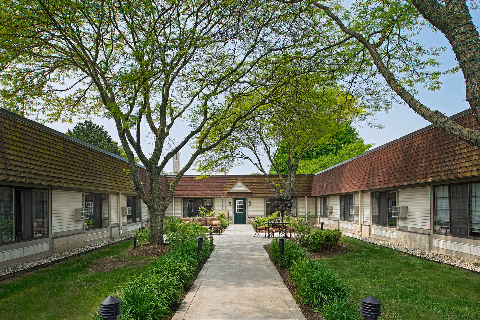Outdoor courtyard area of American House Village of Rochester featuring a concrete walkway lined with green plants and small bushes. The courtyard is surrounded by a single-story building with windows and air conditioning units. There are several trees with green leaves providing shade, and outdoor seating including tables and chairs is visible near the building entrance.