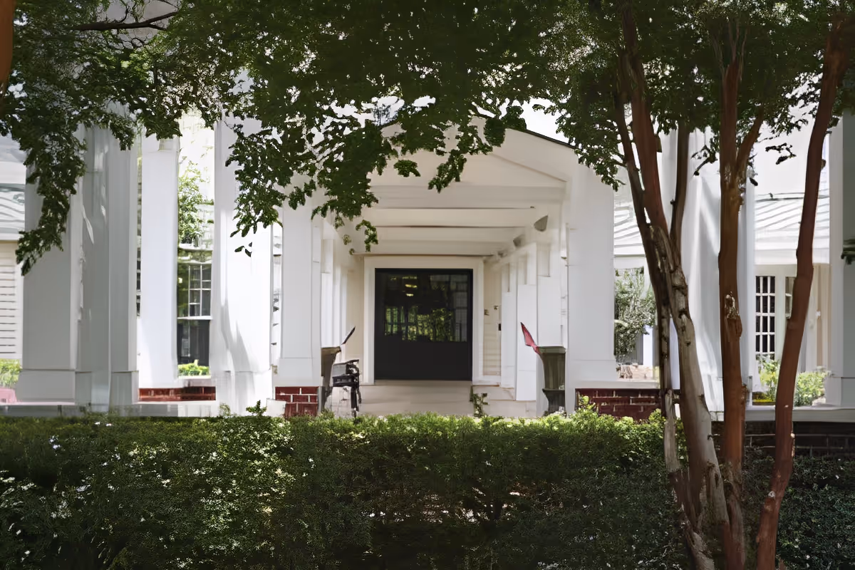Front entrance of a white building with a covered porch supported by columns, surrounded by green bushes and trees.