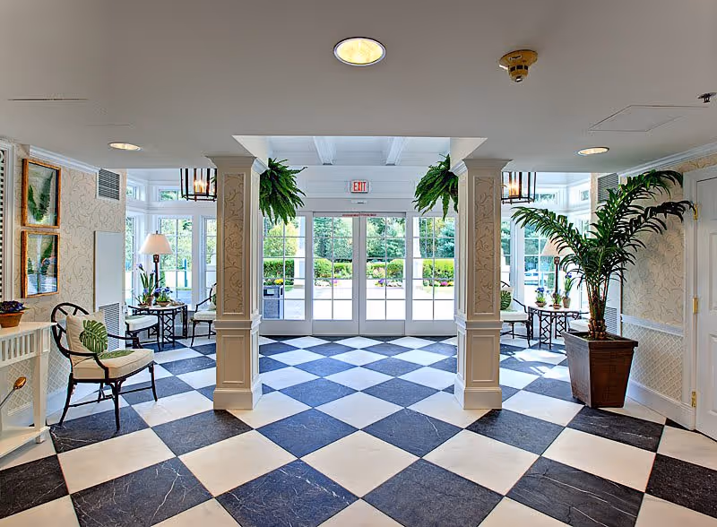 Bright and spacious entrance lobby with black and white checkered floor tiles, white columns, large glass double doors leading outside, potted plants including hanging ferns and a large palm, chairs with cushions, side tables with lamps and decorative items, and framed artwork on the walls.