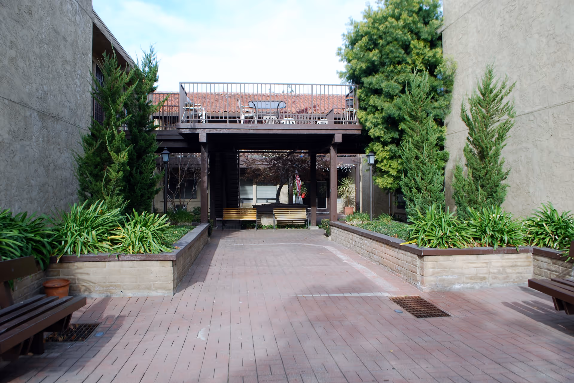 Outdoor courtyard area with brick paving, raised garden beds with green plants and trees, wooden benches along the sides, and a raised deck with tables and chairs above a shaded sitting area.