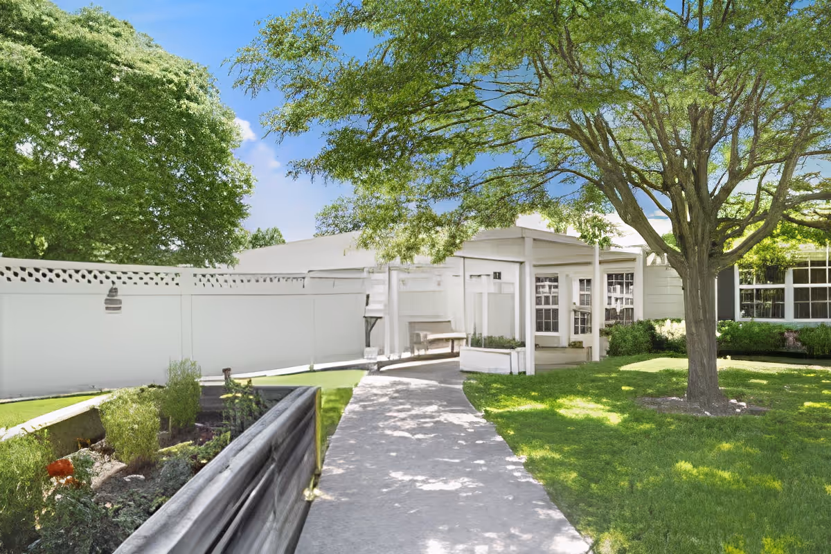 Sunny courtyard with a paved walkway leading to a white building entrance surrounded by lawn, trees, and raised planters.