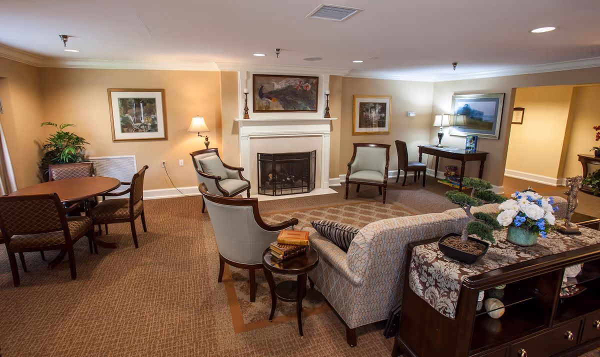 A cozy living room area in a senior living facility featuring a fireplace with artwork above it, several upholstered chairs and a sofa arranged around a small round table with books, a side table with a lamp, framed pictures on the walls, a round dining table with chairs, and decorative plants and flowers on a wooden console table.