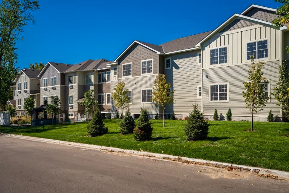 Exterior view of a multi-story senior living facility building with beige siding, multiple windows, and a well-maintained green lawn with small trees and shrubs under a clear blue sky.