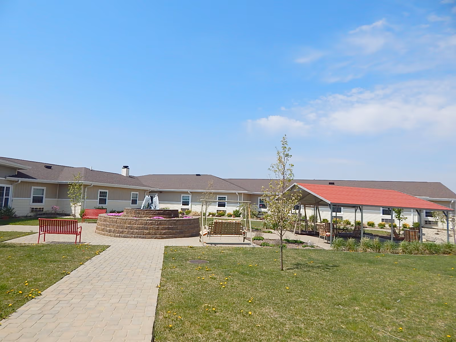 Outdoor courtyard area of a senior living facility with a paved walkway leading to a circular stone fountain surrounded by flowers. There are red benches, a wooden swing, and a covered seating area with a red roof. The building with beige siding and multiple windows surrounds the courtyard under a clear blue sky.