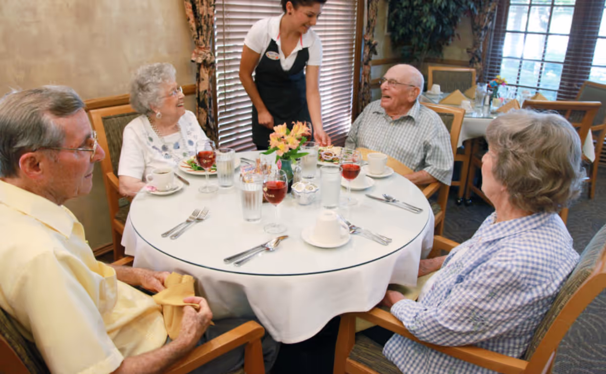 Four elderly residents sit around a round dining table while a staff member serves them in a dining room.
