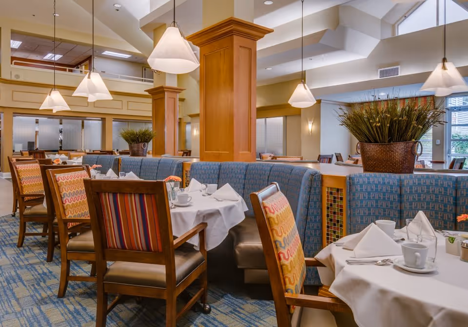 A bright and spacious dining area with tables covered in white tablecloths, set with cups, glasses, napkins, and silverware. The seating includes wooden chairs with colorful patterned cushions and a curved blue upholstered bench. Large pendant lights hang from the ceiling, and there are decorative plants placed on the tables and around the room. The room has high ceilings with skylights and large windows allowing natural light to enter.