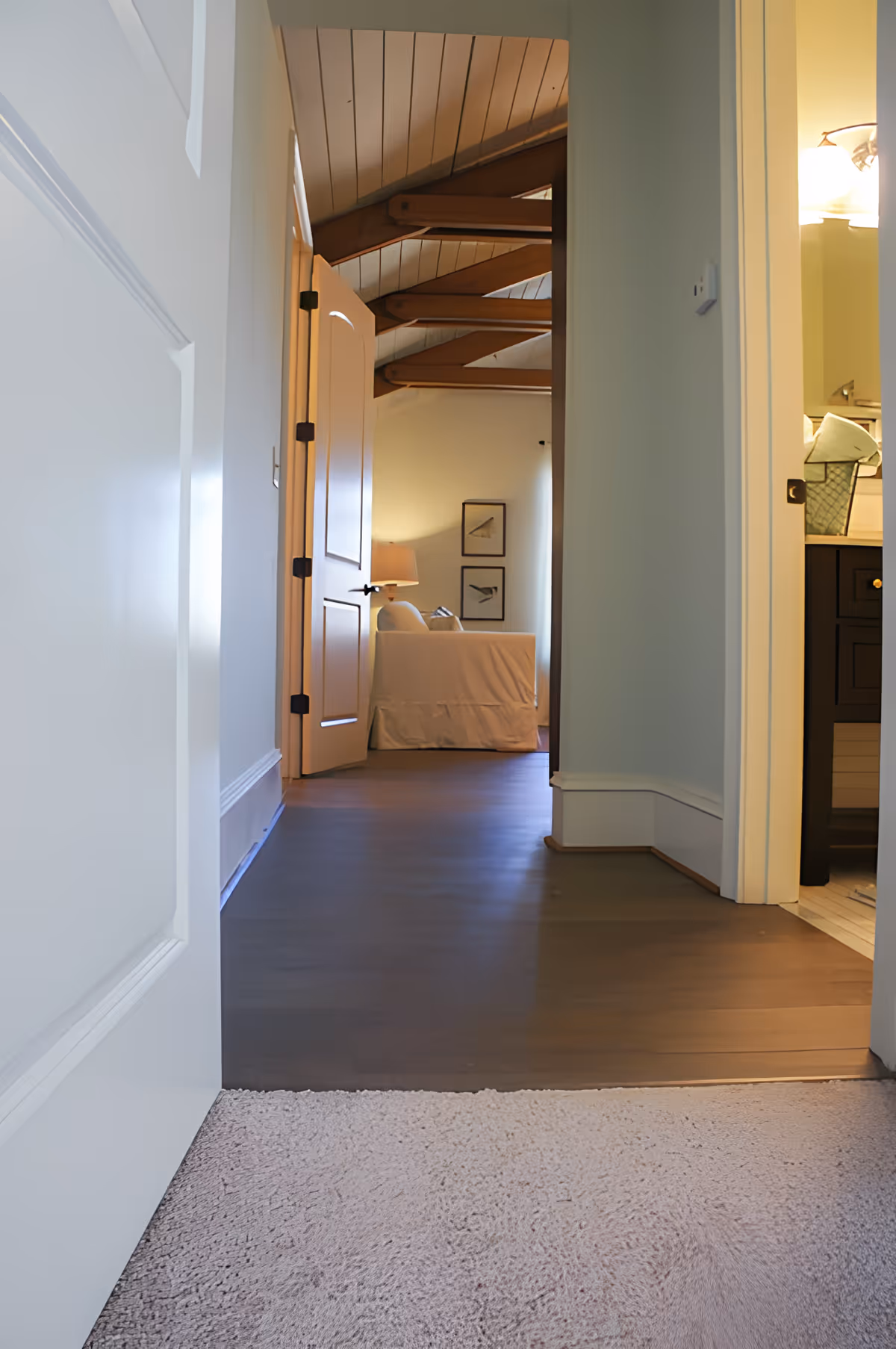 View from a carpeted hallway into a room with wooden flooring, an open door, a white sofa, a lamp, and framed pictures on the wall. The ceiling has exposed wooden beams. To the right, part of a bathroom vanity with a sink and mirror is visible.