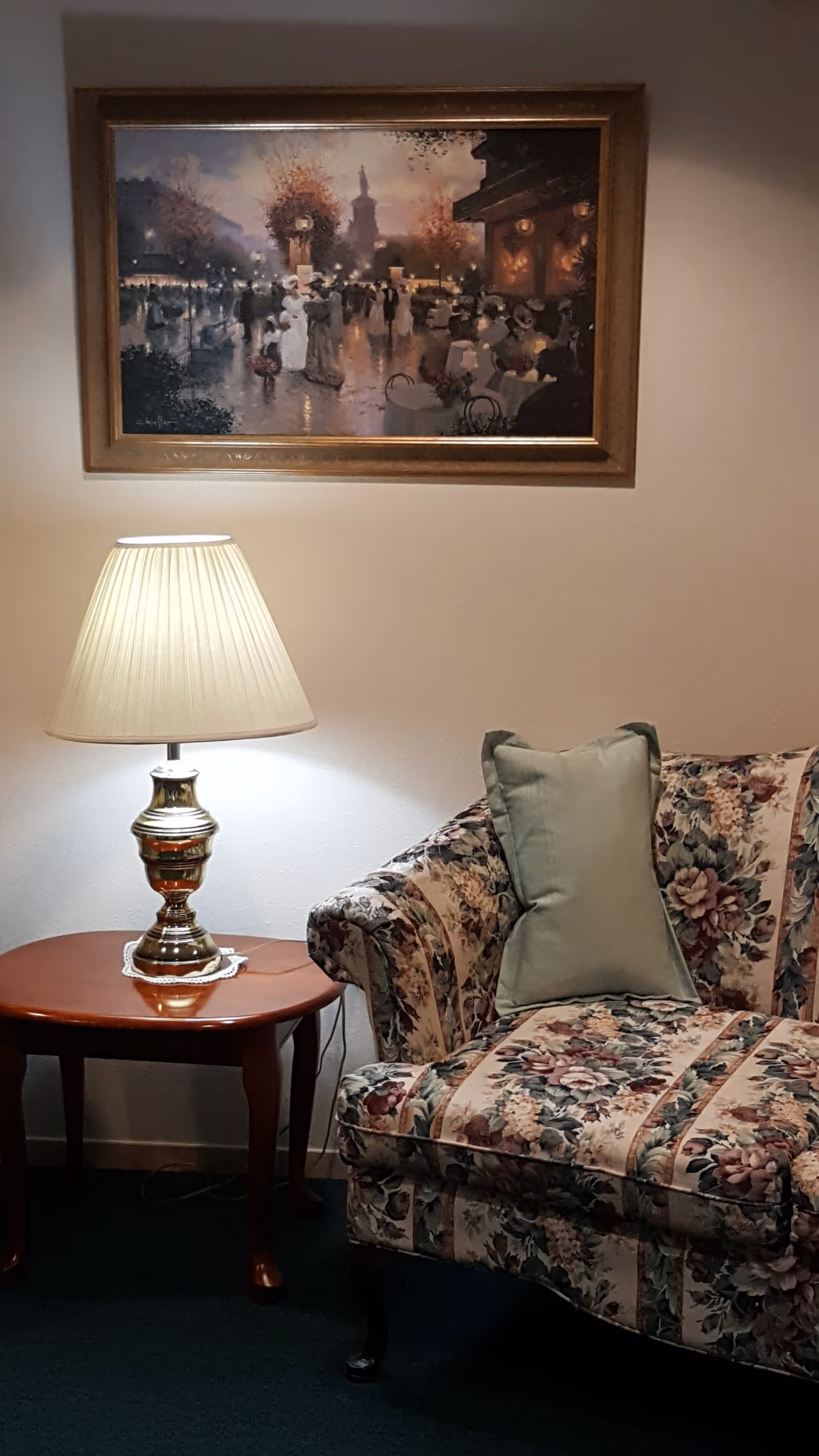 Floral-upholstered couch with a green throw pillow next to a wooden side table holding a lit lamp and a framed painting on the wall above.