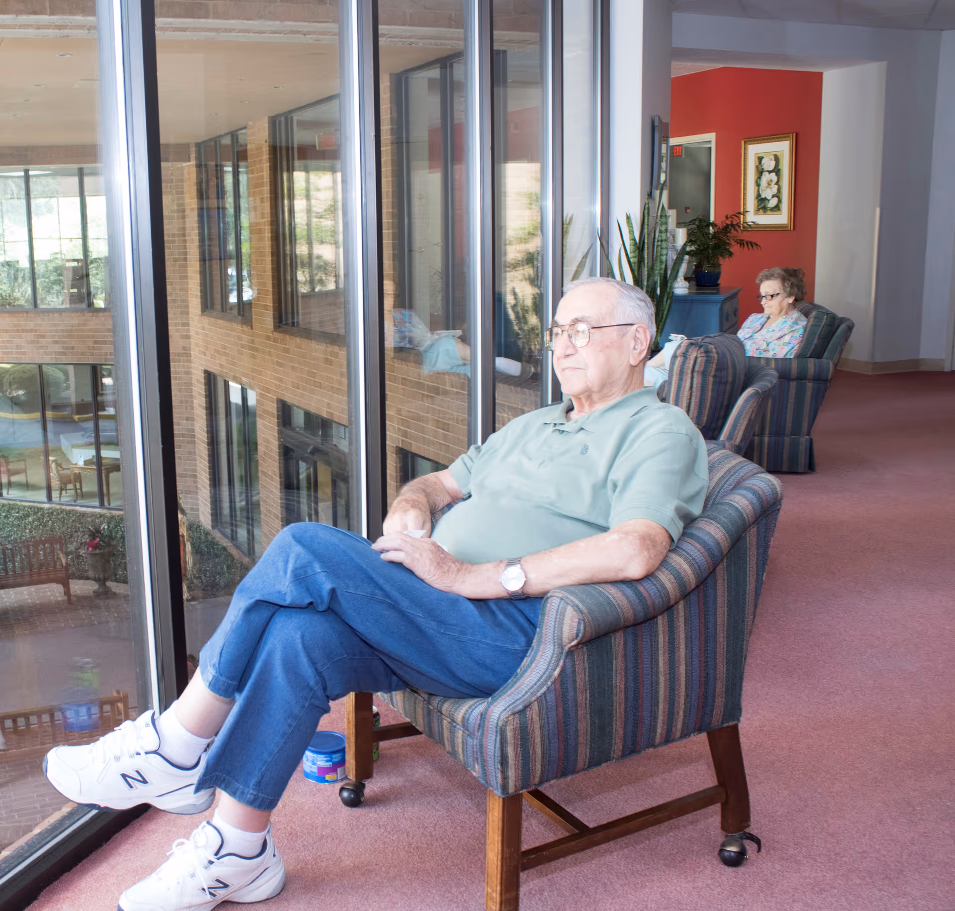 An elderly man wearing glasses, a green polo shirt, blue jeans, and white sneakers sits in a striped armchair by a large window inside a building. Behind him, an elderly woman sits in another armchair near a red wall with a framed floral picture and some plants. The window shows a view of an indoor courtyard with benches and tables.