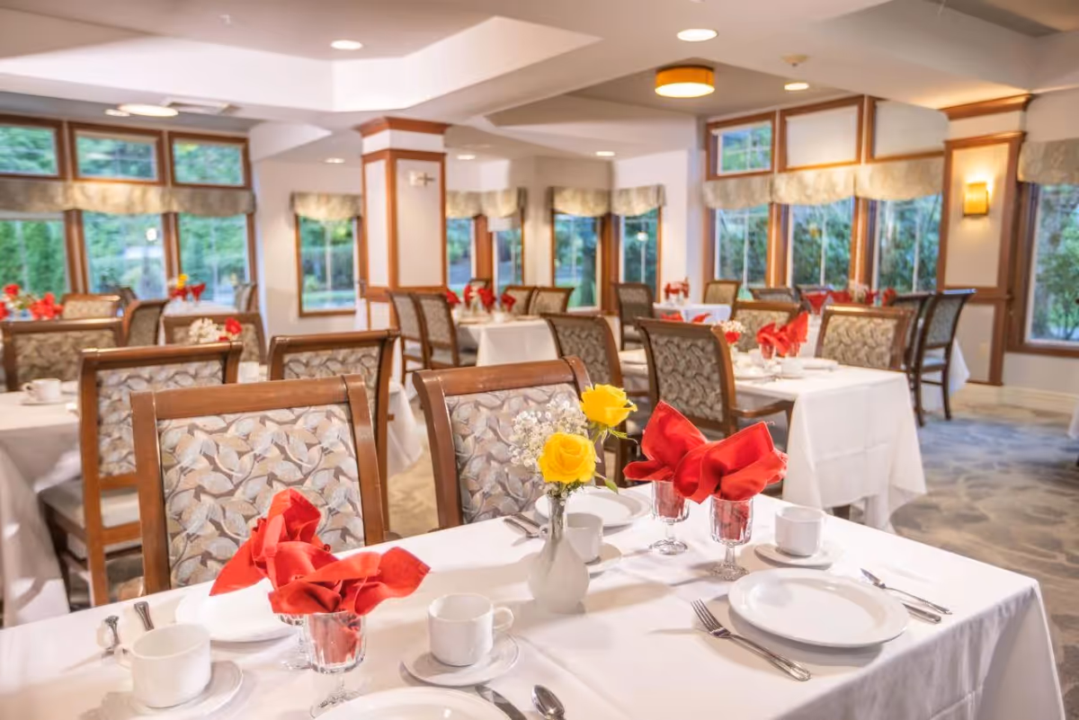A bright and elegant dining room with multiple tables covered in white tablecloths, each set with white plates, cups, silverware, and red folded napkins in glasses. A small vase with yellow roses and white baby's breath flowers is on the nearest table. The room has large windows with beige valances, wooden trim, and soft lighting.