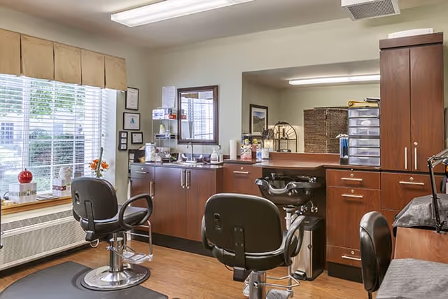 Interior view of a hair salon area in a senior living facility with two black salon chairs, wooden cabinets, a large mirror, and a window with blinds and a beige valance. Various hair care products and supplies are placed on the counter and shelves.