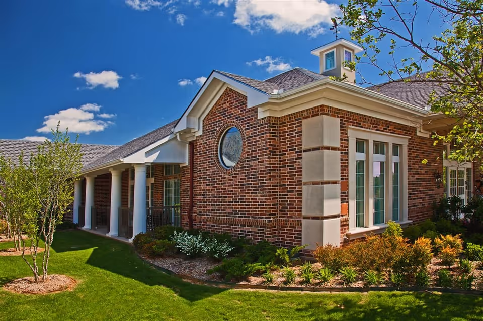 Exterior view of a brick building with white columns and a well-maintained garden under a blue sky with scattered clouds.