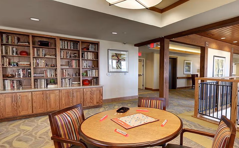 Interior common room with a round game table and chairs, bookshelves and seating in a senior living facility.