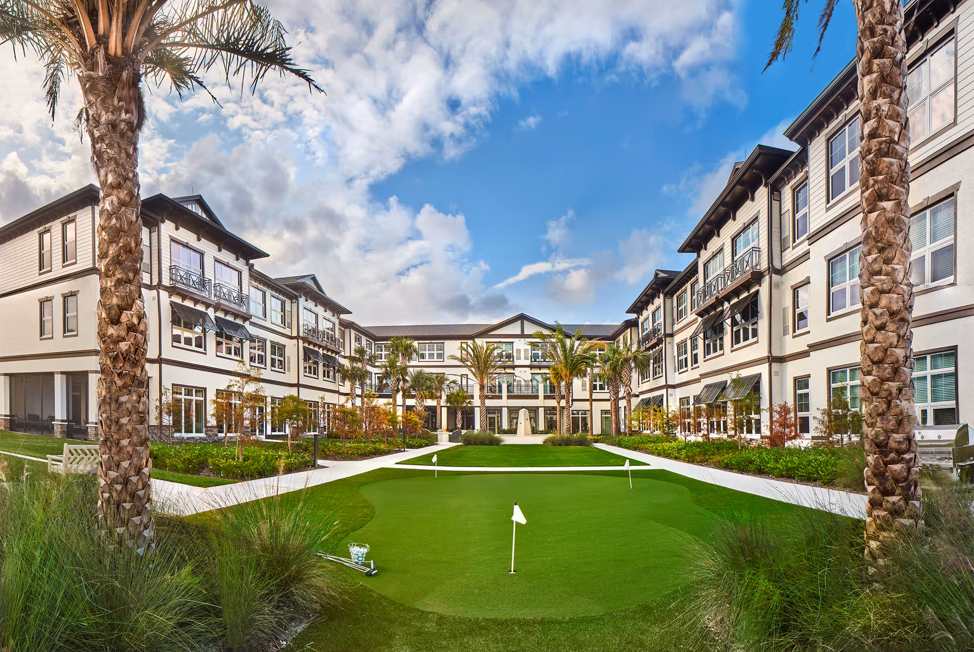 Outdoor courtyard area of a senior living facility with a putting green, palm trees, landscaped gardens, and a three-story building surrounding the courtyard under a partly cloudy sky.