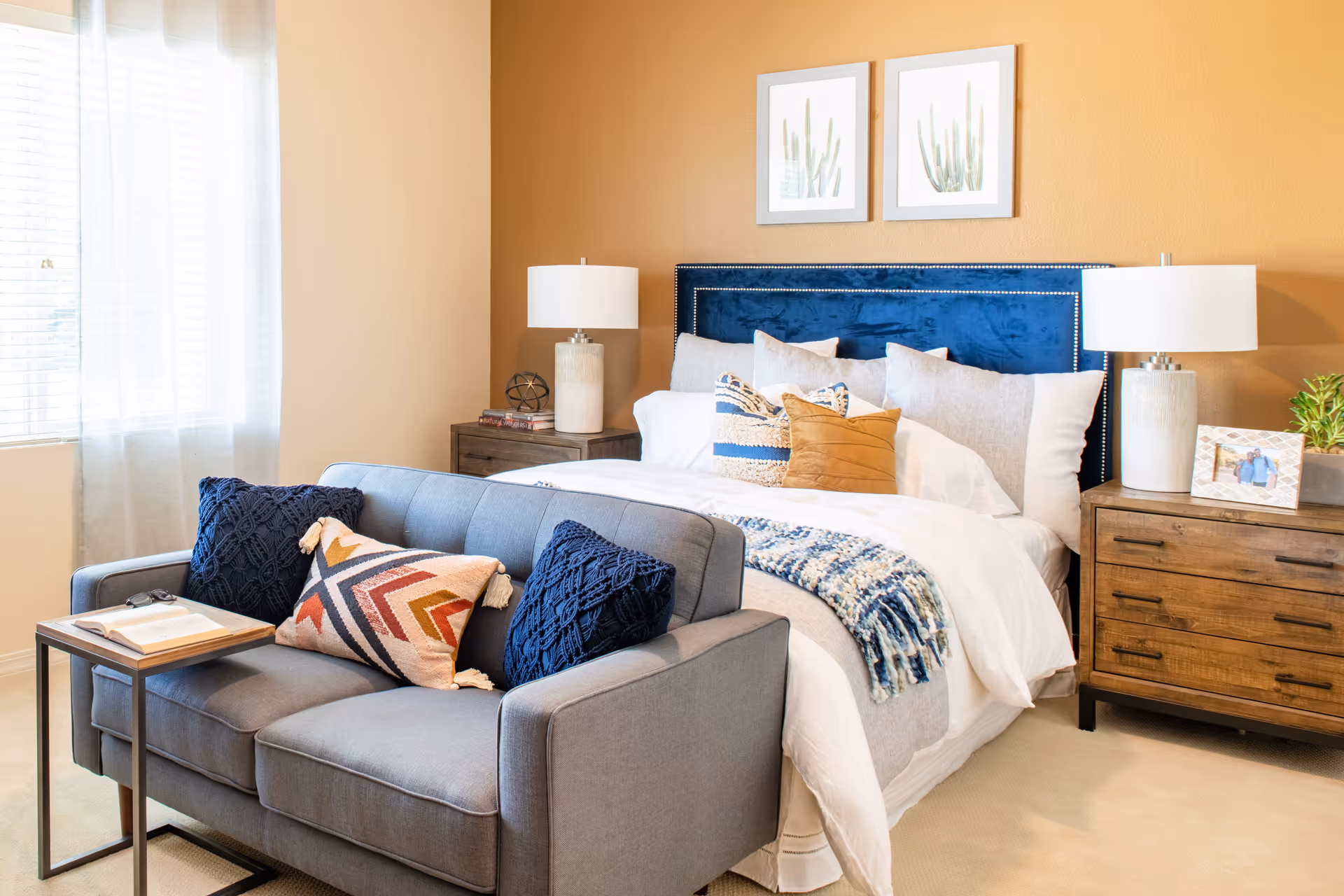 Sunlit bedroom with a blue upholstered headboard, white bedding, a gray loveseat at the foot of the bed, and wooden nightstands with lamps.