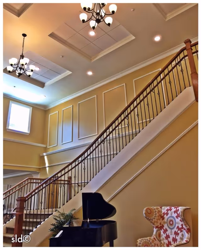 Sunlit two-story interior lobby with a sweeping staircase, chandeliers, a black grand piano and a patterned armchair.