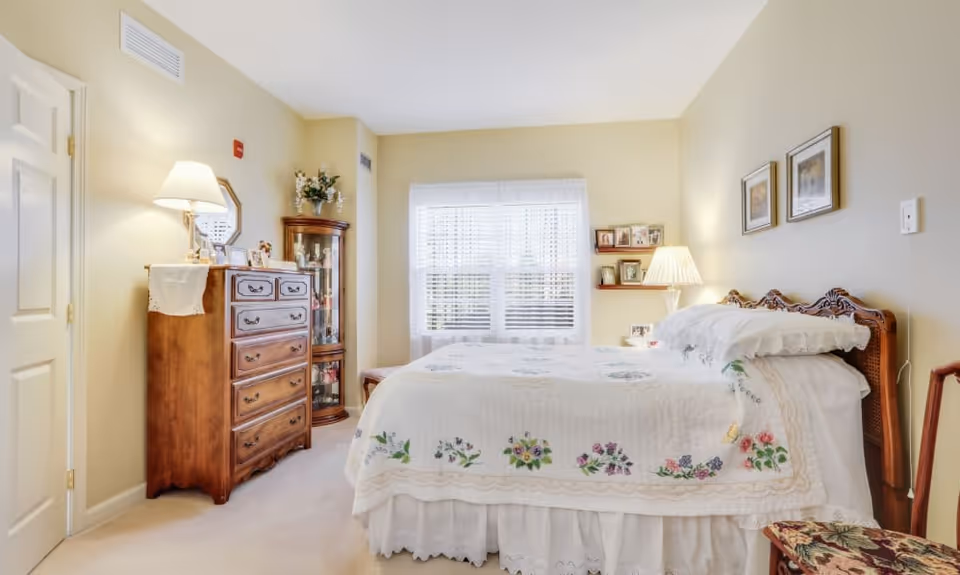 A cozy bedroom with a large bed covered in a white quilt with floral embroidery. The room has beige walls and carpet, a wooden dresser with a lamp and decorative items, a corner glass display cabinet, a window with white blinds and sheer curtains, and framed pictures on the wall. There is also a small wooden chair with a floral cushion.
