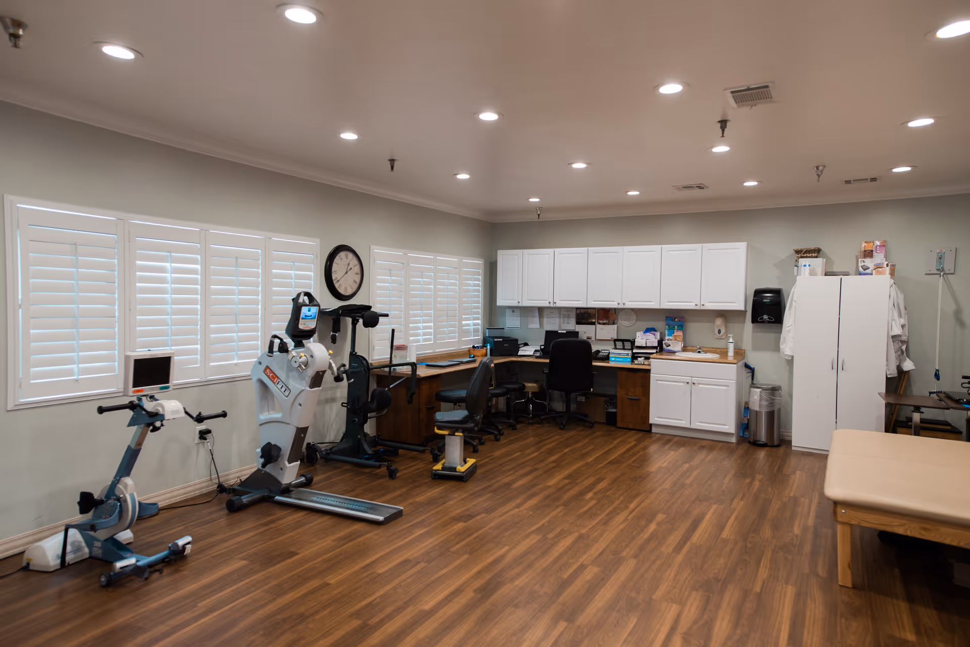 A spacious room with exercise equipment including stationary bikes, a desk with office chairs, white cabinets mounted on the wall, and a treatment table. The room has wooden flooring, multiple windows with white shutters, and ceiling lights.